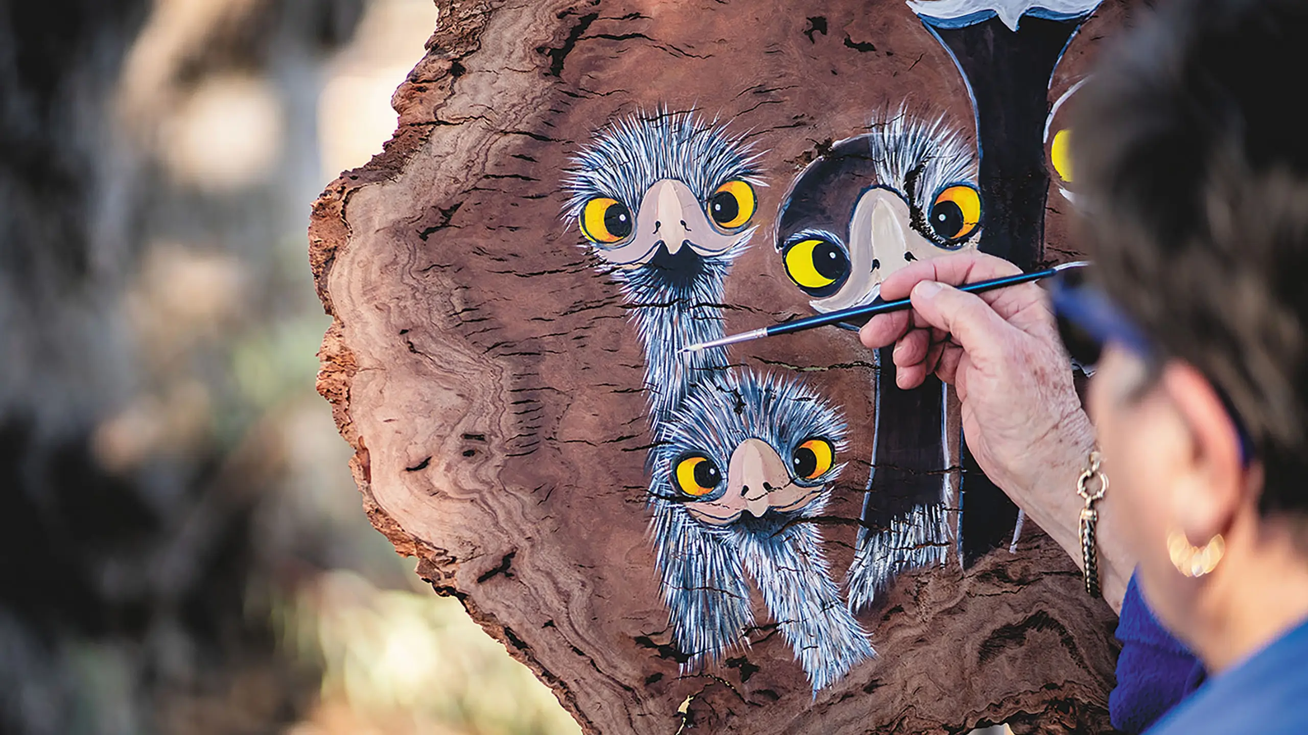 An artist wearing a blue shirt and glasses, on the right side, holding a paintbrush and creating emu art on recovered timber. The timber features three emus.