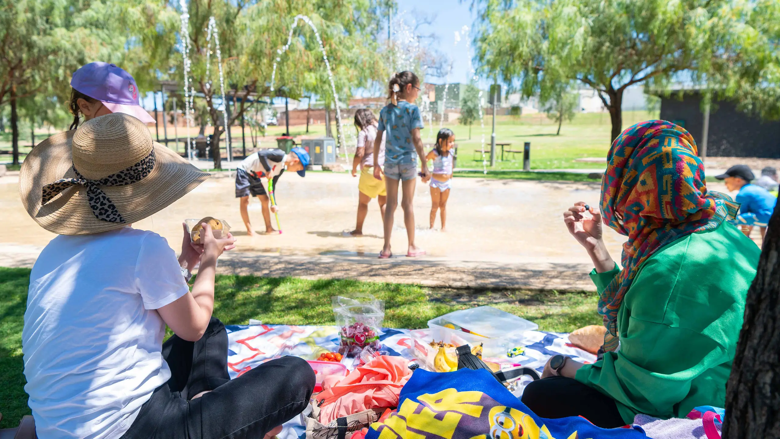 Two women sitting on a picnic mat with food at a water splash park, watching their children play. One woman is wearing a hat and the other is wearing a head scarf.