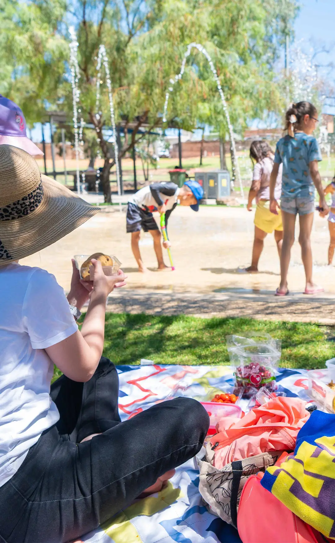 Children playing at a water splash park. A woman is sitting on a picnic mat, holding cookies.