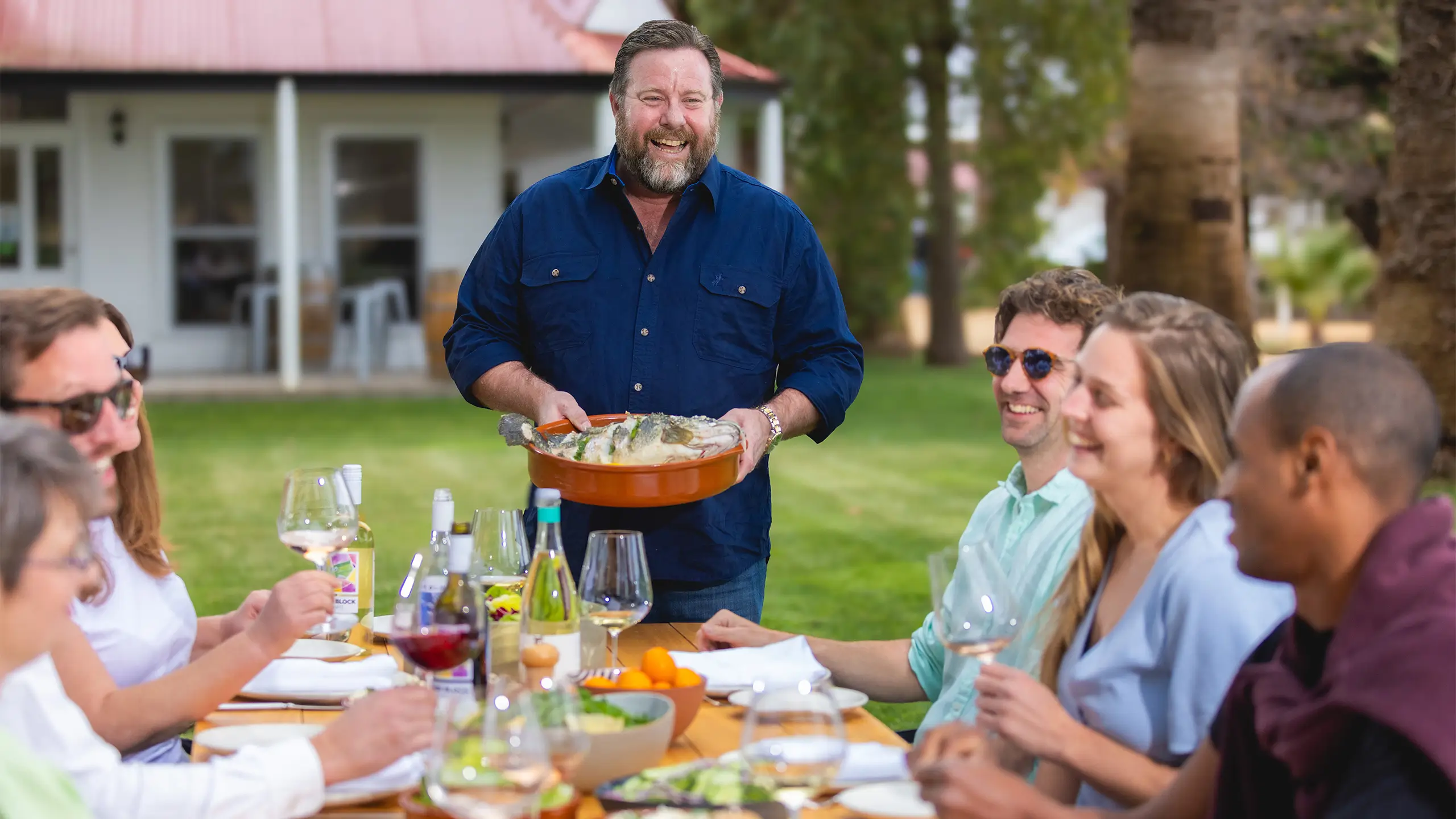 A happy man holding a dish of food in his hand at a table with people smiling and enjoying Mildura cuisine.