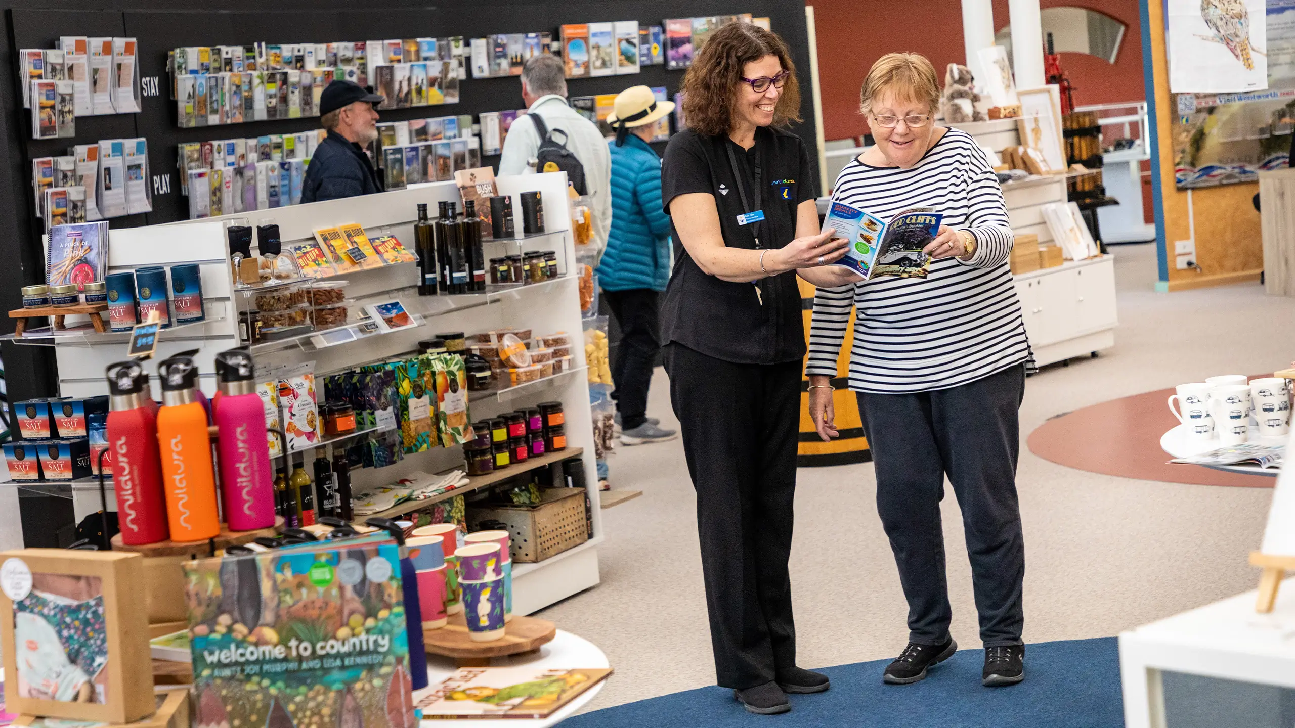 A staff member offering customer service to an elderly female visitor at the Mildura Visitor Information Centre. More visitors are visible in the background, and local merchandise is displayed.