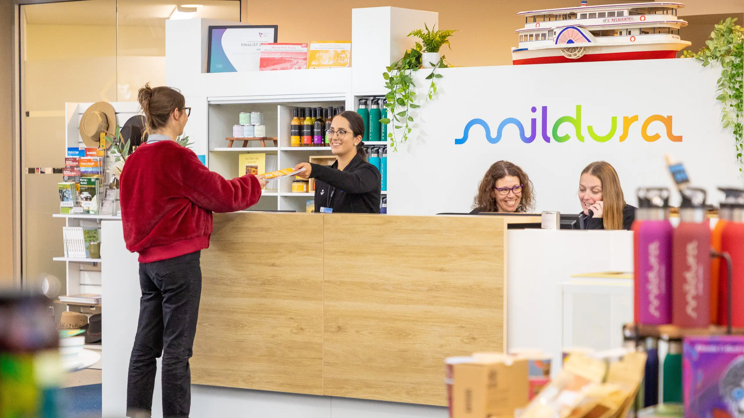 A staff member at the helpdesk handing an information pack to a visitor at the Mildura Visitor Information Centre. Two additional staff members are visible, one on a phone call and the other smiling.